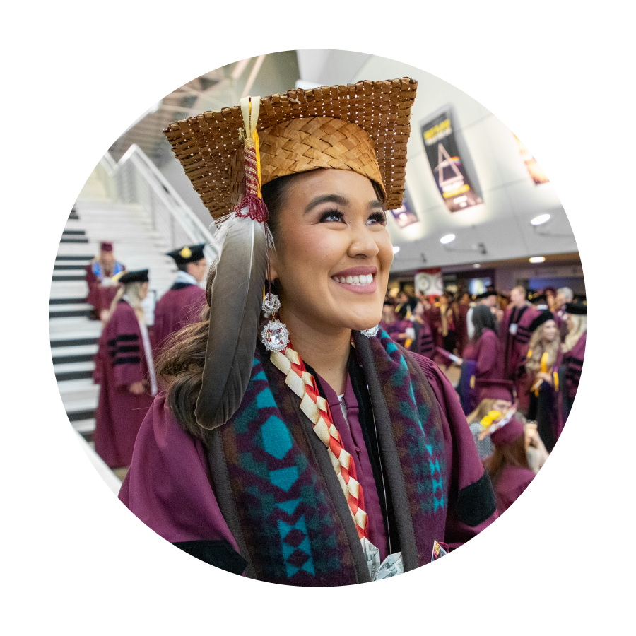 Student smiling in maroon regalia and brown woven cap looking up