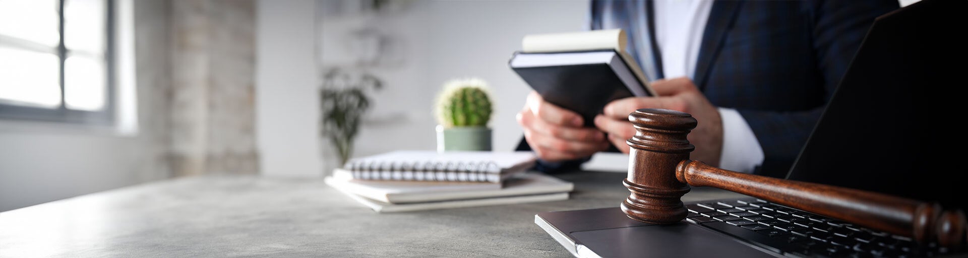 View of two hands holding legal papers while sitting at a desk. Their is a cactus on the desk and a gavel on top of a silver laptop to the side of the person. 