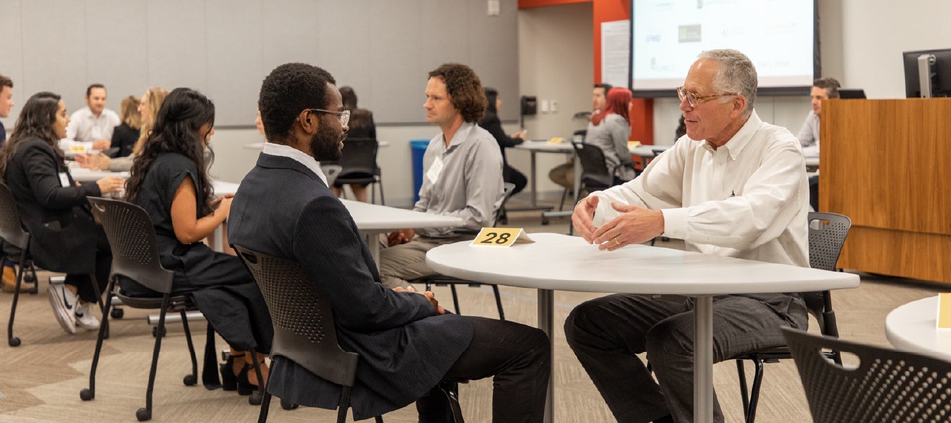 A student dressing in a suit sitting at a table talking to an older man who is speaking and holding his hands out.