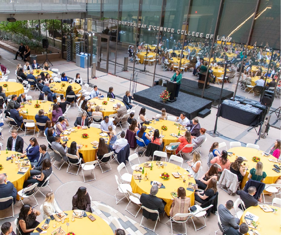 Arial view of students and faculty sitting at tables with yellow tablecloths, eating while attending the Graduation dinner. 