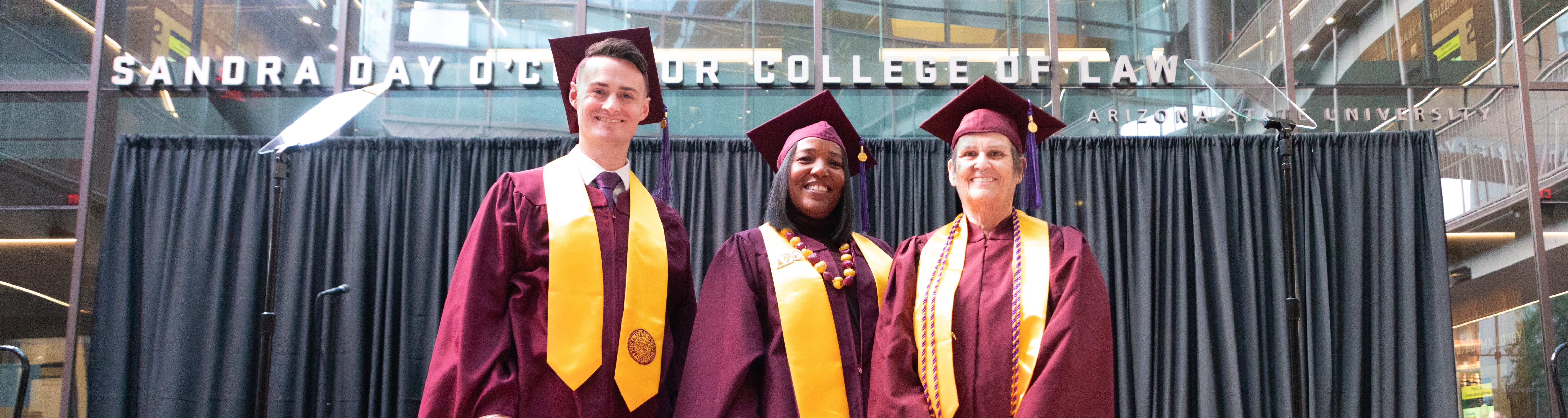 Three smiling Master of Legal Studies students standing on the stage in front of BCLS at the Fall 2023 Convocation ceremony.