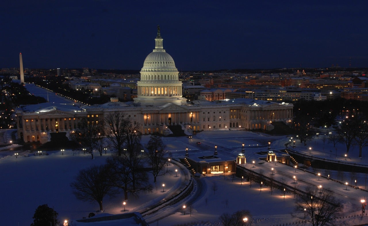 United States Capitol at night covered in snow. The building is lit up with bright white lights in a blue dark dusk sky.