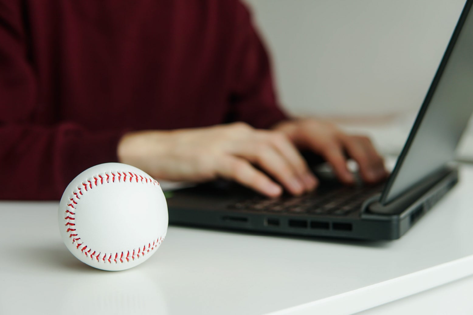 A man checking baseball stats on a laptop. A white baseball sits next to him on the table.