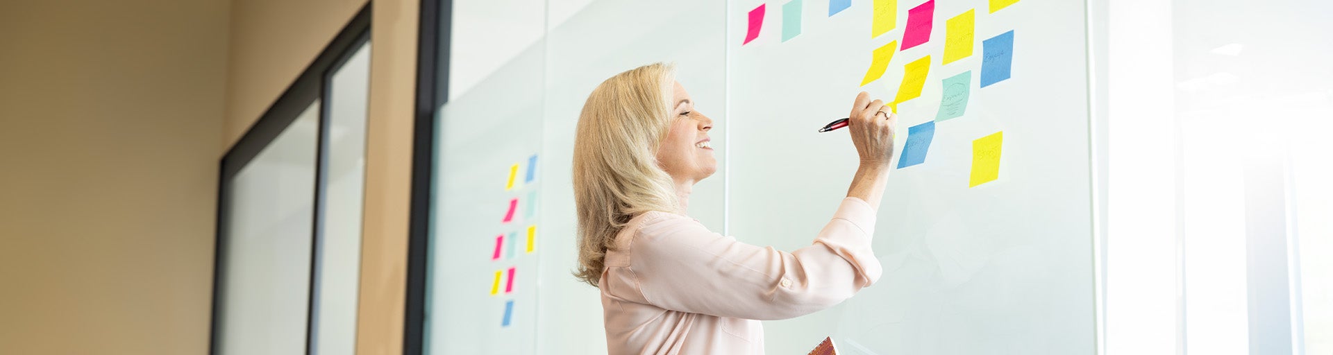 Woman writing on colorful post-it notes that are stuck to a opaque wall.