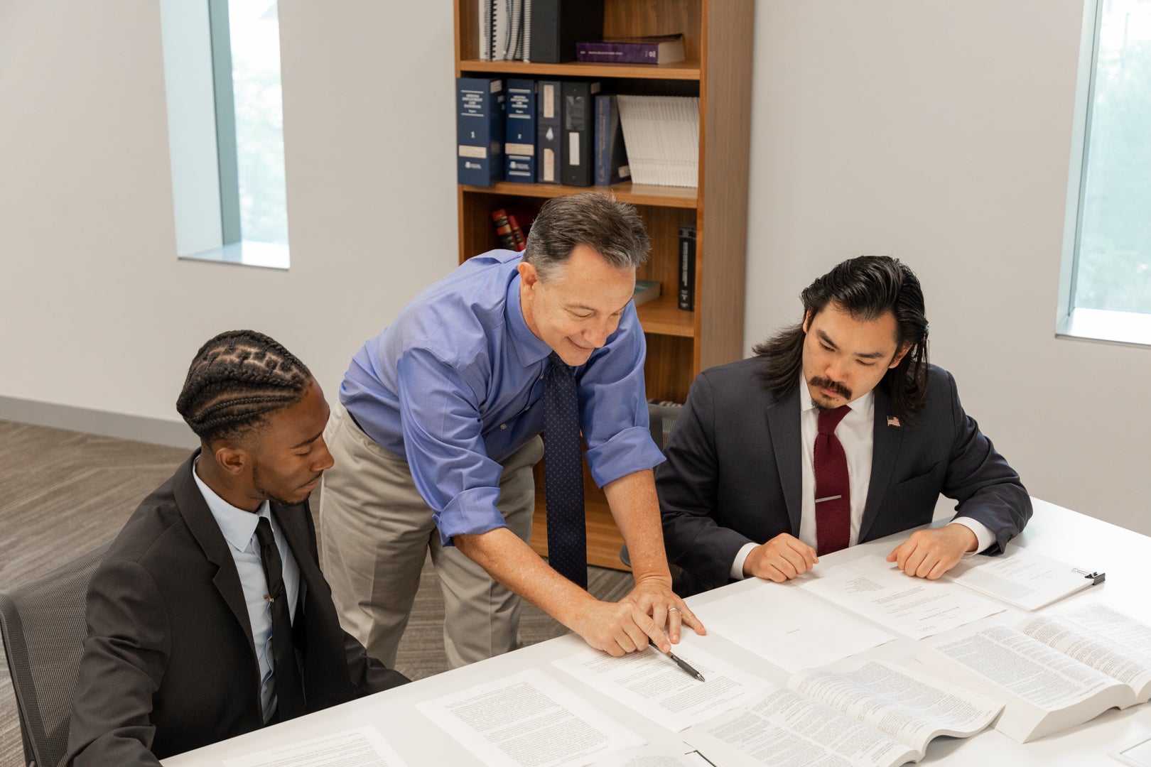 Professor Hindshaw pointing to a page with a pen while two students look over his shoulder. 