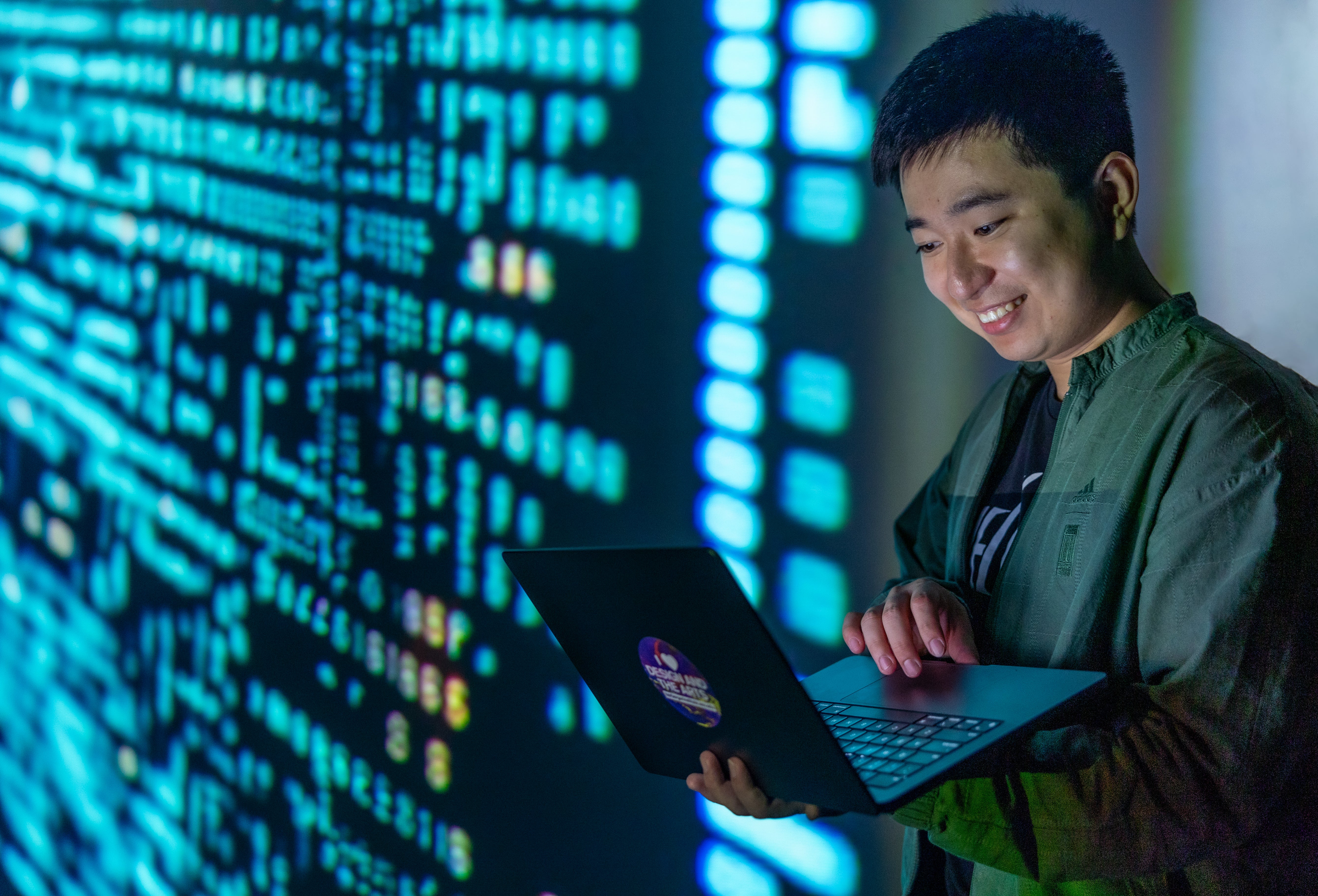 ASU student standing and holding their laptop while blue code and characters are projected on the wall next to him. 