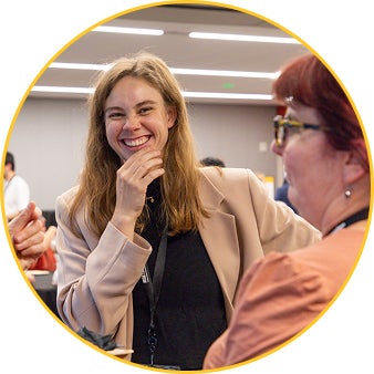 A student smiles broadly, with their hand partially covering their mouth, talking with others in a bright indoor setting.