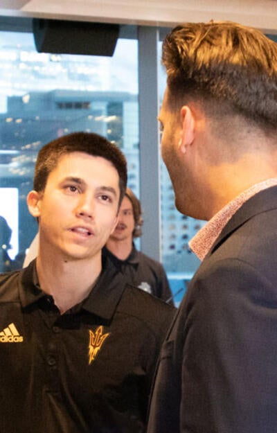 A male student wearing a ASU Sun Devils polo shirt talks to another male student in the foreground