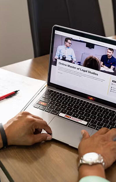 Two hands type at a laptop with a webpage visible that reads "Online Master of Legal Studies"