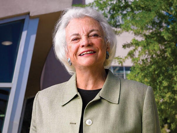 The. Honorable Sandra Day O’Connor standing and smiling in front of a building a tree. She is wearing a green blazer.