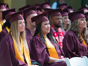 A group of graduates in maroon robes sits in folding chairs during a ceremony.