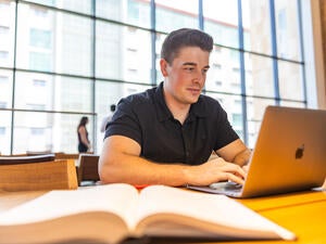 Male student sits at a table in the Sandra Day O'Connor College of Law Reading Room working on a laptop with an open book in front of him.