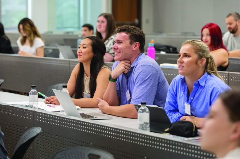 Three students smiling while sitting in lecture hall during orientation watching the front of the classroom