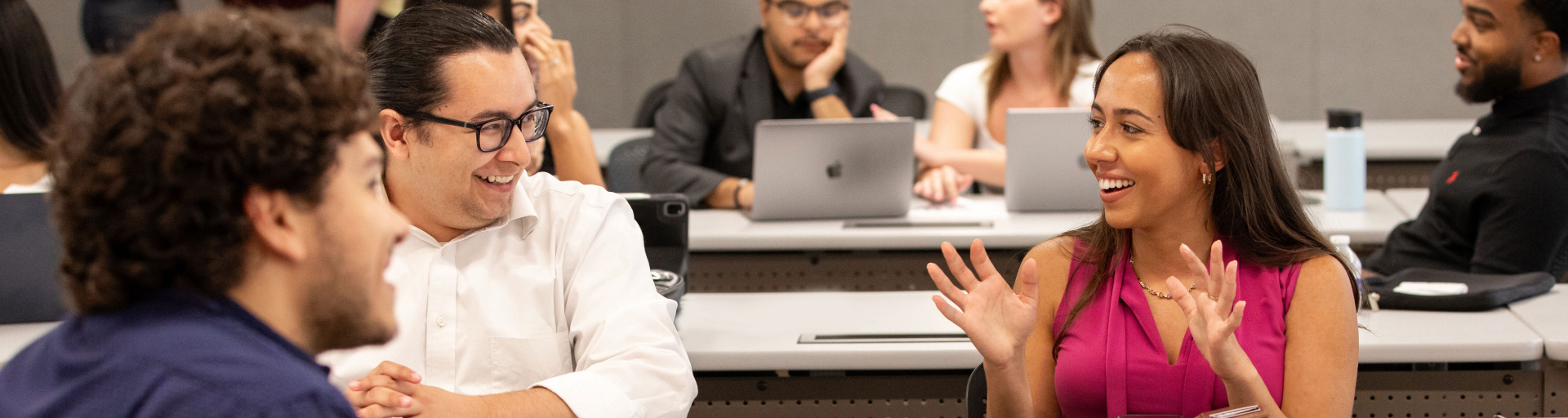 Three students talking to each other while sitting at a long white desk in an ASU Law classroom, surrounded by other students.