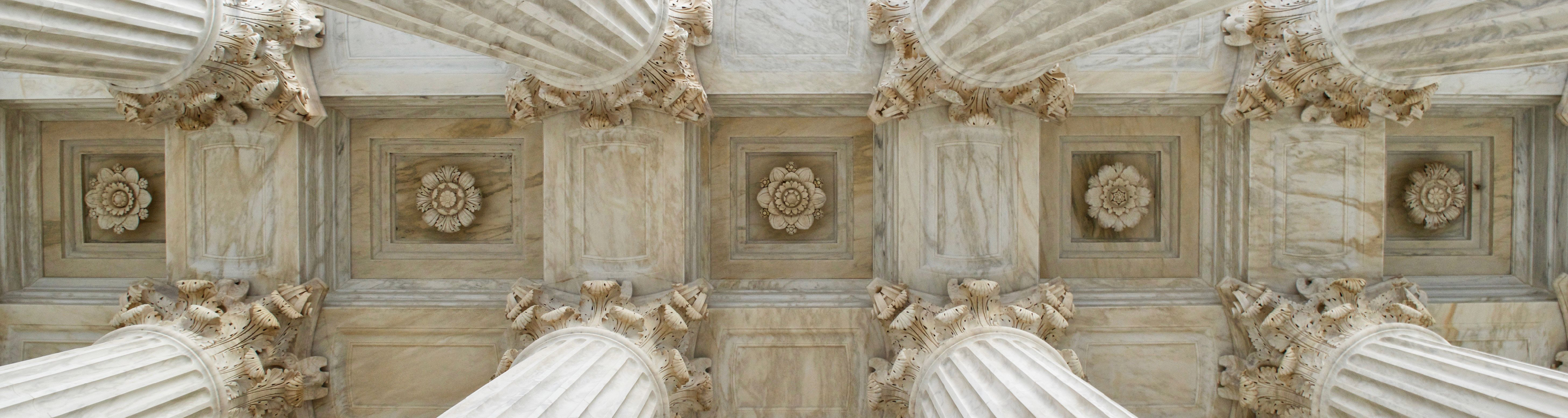 View of the ceiling of a ivory government building from below. The ceiling consists of floral patterns, squares, and large ivory decorative columns.