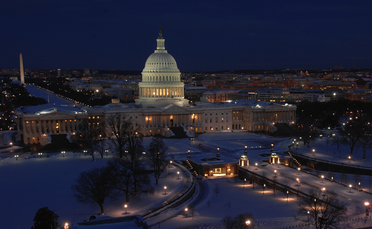United States Capitol at night covered in snow. The building is lit up with bright white lights in a blue dark dusk sky.